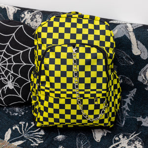 The CHOK yellow checker vegan backpack is sat on a spooky halloween blanket next to a spiderweb cushion facing forward. The backpack is yellow and black all over checkerboard with two front black zip pockets, two side pockets and a silver decorative chain going across the front.