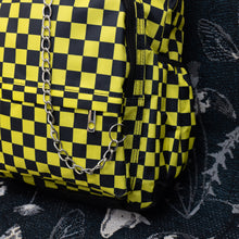 Load image into Gallery viewer, Close up of the CHOK yellow checker vegan backpack sat on a spooky halloween blanket next to a spiderweb cushion facing forward. The backpack is yellow and black all over checkerboard with two front black zip pockets, two side pockets and a silver decorative chain going across the front.
