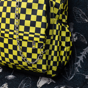 Close up of the CHOK yellow checker vegan backpack sat on a spooky halloween blanket next to a spiderweb cushion facing forward. The backpack is yellow and black all over checkerboard with two front black zip pockets, two side pockets and a silver decorative chain going across the front.
