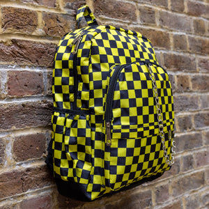 The CHOK yellow checker vegan backpack hanging up on a brick wall outside facing right. The backpack is yellow and black all over checkerboard with two front black zip pockets, two side pockets and a silver decorative chain going across the front.