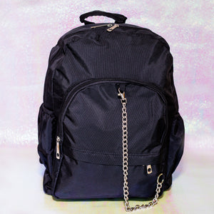 The black nylon vegan backpack with chain on an iridescent background. The bag is facing forward to highlight the two front zip pockets, two side pockets, double zip main compartment, top handle and detachable silver chain.