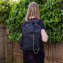 Load image into Gallery viewer, Jack is stood outside in a garden area wearing the black nylon vegan backpack with chain on his back whilst facing away from the camera. The photo is cropped from the thighs up. The backpack is all black with two front zip pockets, two side pockets and a silver decorative chain across the front to the side.
