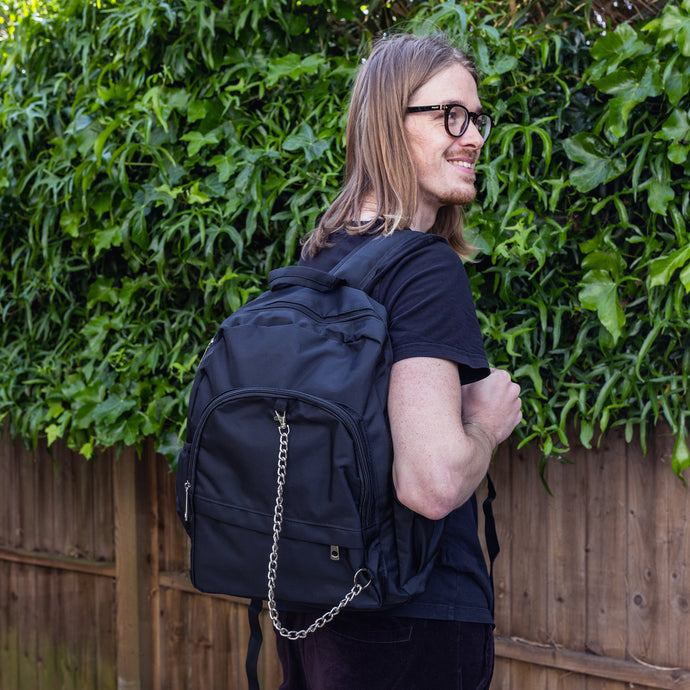 Jack is stood outside in a garden area wearing the black nylon vegan backpack with chain on his back whilst smiling looking right. The photo is cropped from the thighs up. The backpack is all black with two front zip pockets, two side pockets and a silver decorative chain across the front to the side.