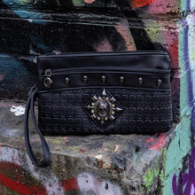 Load image into Gallery viewer, The gothx black skull vegan clutch bag on an alternative colourful graffiti brick wall outside. The vegan clutch bag is facing forward to highlight the skull embossed vegan black leather, the crystal stud skull centrepiece, zip pocket and skull studs.
