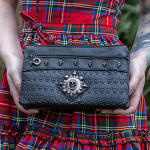 The GothX black skull vegan clutch bag being held by a tattooed model in front of their red tartan dress. The clutch is facing forward to highlight the skull embossed vegan black leather, the crystal stud skull centrepiece, zip pocket and skull studs.
