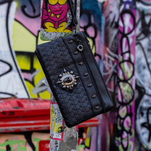 Load image into Gallery viewer, The gothx black skull vegan clutch bag on an alternative colourful graffiti brick wall outside. The vegan clutch bag is facing forward to highlight the skull embossed vegan black leather, the crystal stud skull centrepiece, zip pocket and skull studs.
