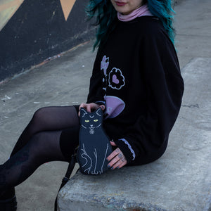Model with blue hair wearing a hello kitty outfit holding the gothx pagan black cat vegan shoulder bag next to them. The bag is facing forward to highlight the embroidery details including a pentagram collar, yellow eyes and paws.