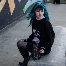 Load image into Gallery viewer, Model with blue hair wearing a hello kitty outfit holding the gothx pagan black cat vegan shoulder bag next to them. The bag is facing forward to highlight the embroidery details including a pentagram collar, yellow eyes and paws.
