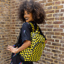 Load image into Gallery viewer, Cindy is stood outside in front of a brick wall wearing the CHOK yellow checker vegan backpack on her back whilst looking over her shoulder to camera. The backpack is yellow and black checkerboard all over print with two front zip pockets, two side pockets and a silver decorative chain going across the front.
