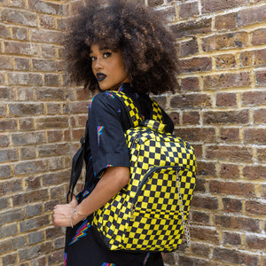 Cindy is stood outside in front of a brick wall wearing the CHOK yellow checker vegan backpack on her back whilst looking over her shoulder to camera. The backpack is yellow and black checkerboard all over print with two front zip pockets, two side pockets and a silver decorative chain going across the front.