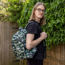 Load image into Gallery viewer, Jack is stood in a garden area modelling the classic camouflage camo vegan backpack. The bag is facing towards the camera to highlight the front camo print, two front zip pockets, two elastic side pockets, detachable silver chain and top handle.
