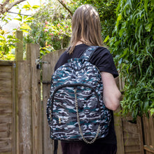 Load image into Gallery viewer, Jack is stood in a garden area modelling the forest camouflage camo vegan backpack. The bag is facing towards the camera to highlight the front camo print, two front zip pockets, two elastic side pockets, detachable silver chain and top handle.
