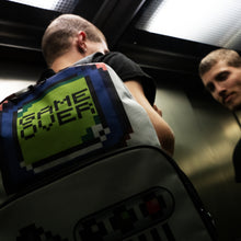 Load image into Gallery viewer, A man in a dark lift wearing the retro white game over vegan backpack. The backpack features a 90s inspired gameboy print with buttons and a screen saying game over.
