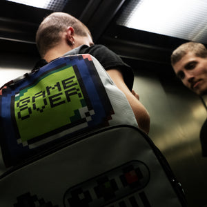A man in a dark lift wearing the retro white game over vegan backpack. The backpack features a 90s inspired gameboy print with buttons and a screen saying game over.