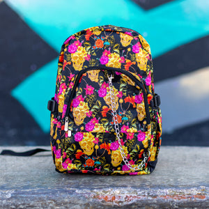 The floral gold skull nylon chain vegan backpack sat on a skatepark bench. The bag is facing forward to highlight the flowers and skulls print, two zipped pockets, two elastic side pockets and detachable decorative silver chain.
