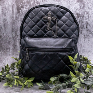 The GothX quilted cross vegan mini backpack on a grey studio background with leaves surrounding it. Quilted front detailing with a studded cross with hanging chain appliqué and zip front pocket. Black vegan leather with gunmetal grey detailing. The bag is facing forward.