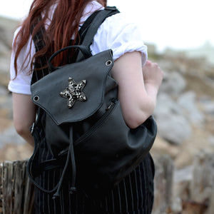 GothX Skull and Star Black Vegan Tassel Tie Backpack shown being held by a model wearing pin stripe dungarees and a white short sleeve top. They are walking away from the camera with the bag on one shoulder whilst their hand rests on the front strap. Bag has adjustable vegan leather straps with tassel tie and metal magnetic clip close with large zipped front pocket.