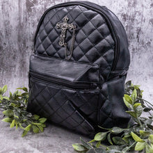 Load image into Gallery viewer, The GothX quilted cross vegan mini backpack on a grey studio background with leaves surrounding it. Quilted front detailing with a studded cross with hanging chain appliqué and zip front pocket. Black vegan leather with gunmetal grey detailing. The bag is angled to the left to show the depth of the bag. 
