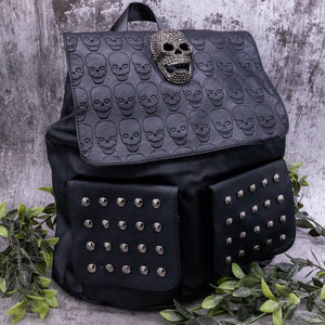 The GothX twin pocket skull vegan backpack on a grey velvet background with leaves surrounding it. The bag is facing forward to highlight the diamante effect skull, skull embossed vegan leather front flap, tassel tie cords and two silver studded front pockets.
