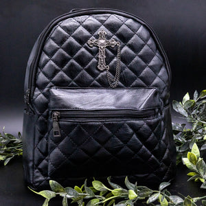 The GothX quilted cross vegan mini backpack on a black studio background with black roses and leaves surrounding it. Quilted front detailing with a studded cross with hanging chain appliqué and zip front pocket. Black vegan leather with gunmetal grey detailing. The bag is facing forward to show off the front details.