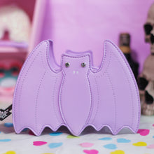 Load image into Gallery viewer, The GothX Pastel Lilac Purple Bat Vegan Shoulder Bag on a pastel purple background with multicoloured confetti, pastel pink coffin shelving, skulls and black skull bottles surrounding it. The bag is facing forward to highlight the embroidered detailing and crystal eyes.
