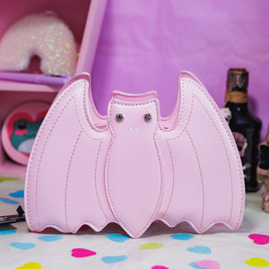 The GothX LIMITED EDITION Pastel Pink Bat Vegan Shoulder Bag on a pastel purple background with multicoloured confetti, pastel pink coffin shelving and black skull bottles surrounding it. The bag is facing forward to highlight the embroidered detailing and crystal eyes.