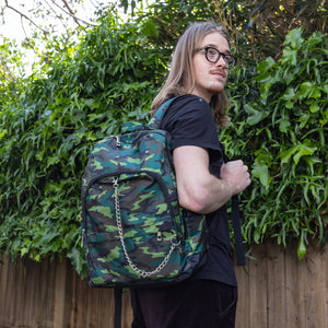 Jack is stood in a garden area modelling the jungle camouflage camo vegan backpack. The bag is facing towards the camera to highlight the front camo print, two front zip pockets, two elastic side pockets, detachable silver chain and top handle.