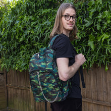 Load image into Gallery viewer, Jack is stood in a garden area modelling the jungle camouflage camo vegan backpack. The bag is facing towards the camera to highlight the front camo print, two front zip pockets, two elastic side pockets, detachable silver chain and top handle.
