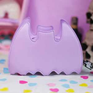 The GothX LIMITED EDITION Pastel Lilac Purple Bat Vegan Shoulder Bag on a pastel purple background with multicoloured confetti, pastel pink coffin shelving and black skull bottles surrounding it. The bag is facing away to highlight the plain pastel purple back.