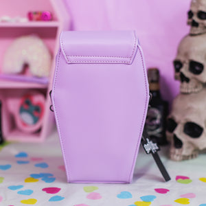 The GothX Pastel Lilac Mini Coffin Vegan Cross Body Bag on a pastel purple background with pastel pink coffin shelving, skulls and black faux skull poison bottles in the background. The bag is facing away to highlight the plain pastel purple vegan leather back.