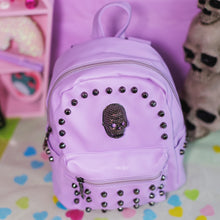 Load image into Gallery viewer, The GothX Pastel Lilac Skull Head Small Studs Vegan Mini Backpack on a pastel purple background with skulls and pastel pink coffin shelf in the background. The bag is facing forward angled right to highlight the dark grey metal stud detailing, diamanté effect skull, main zip compartment, front zip pocket, two side slip pockets and top handle.

