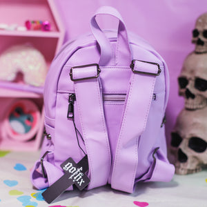 The GothX LIMITED EDITION Pastel Lilac Skull Head Small Studs Vegan Mini Backpack on a pastel purple background with skulls and pastel pink coffin shelf in the background. The bag is facing away to highlight the back zip pocket, adjustable shoulder straps and top handle.