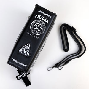 The GothX Ouija Spirit Book Mini Bag sat on a white background. The bag is on its side to highlight the ouija spirit book white printed detailing on the black book bag spine with the 3D planchette stitching. Next to the bag on the right is the adjustable detachable strap. Bag is inspired by witchy style and necromancy.
