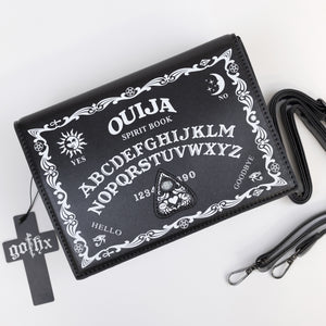 The GothX Ouija Spirit Book Mini Bag sat on a white background. The bag is forward to highlight the ouija spirit book white printed detailing on the black book bag with the 3D planchette stitching. Next to the bag on the right is the adjustable detachable strap. Bag is inspired by witchy style and necromancy.