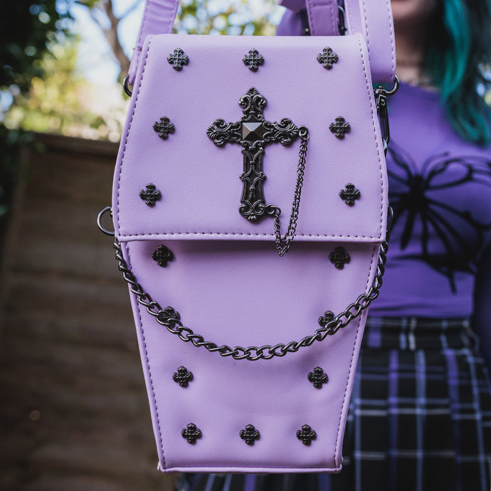 The GothX Pastel Lilac Mini Coffin Vegan Cross Body Bag being held up by a gothic alternative model. The vegan friendly pastel leather bag is facing forward to highlight the cross studs, cross & chain centrepiece, detachable chain and detachable adjustable shoulder strap.
