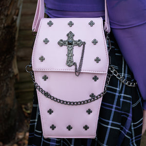 GothX Pastel Pink Mini Coffin Vegan Cross Body Bag being held up by an alternative goth model. The bag is facing forward to highlight the cross studs, cross & chain centrepiece, detachable chain and detachable adjustable shoulder strap.