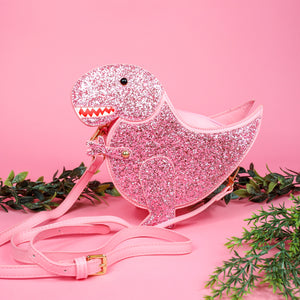 The kawaii pink glitter dino vegan bag on a pink studio background with green foliage. The bag is facing forward to highlight the pastel pink glittered side, dinosaur face, gold metal detailing, detachable strap and moveable arm.