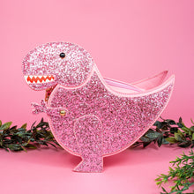 Load image into Gallery viewer, The kawaii pink glitter dino vegan bag on a pink studio background with green foliage. The bag is facing forward to highlight the pastel pink glittered side, dinosaur face, gold metal detailing, detachable strap and moveable arm.
