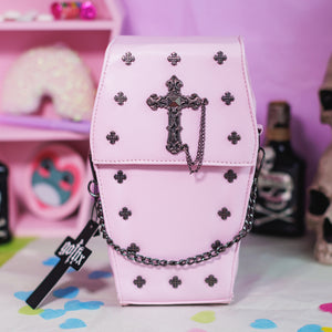 The GothX Pastel Pink Mini Coffin Vegan Cross Body Bag on a pastel purple background with pastel pink coffin shelving, skulls and black faux skull poison bottles in the background. The bag is facing forward to highlight the small cross studs, cross and chain centrepiece and detachable decorative chain.