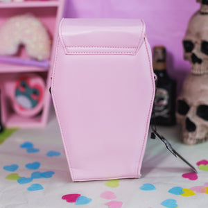 The GothX Pastel Pink Mini Coffin Vegan Cross Body Bag on a pastel purple background with pastel pink coffin shelving, skulls and black faux skull poison bottles in the background. The bag is facing away to highlight the plain pastel pink vegan leather back.