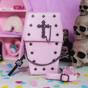 The GothX Pastel Pink Mini Coffin Vegan Cross Body Bag  on a pastel purple background with pastel pink coffin shelving, skulls and black faux skull poison bottles in the background. The bag is facing forward to highlight the cross studs, cross & chain centrepiece, detachable chain and detachable adjustable shoulder strap.