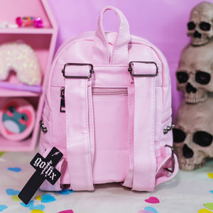 The GothX Pastel Pink Skull Head Small Studs Vegan Mini Backpack on a pastel purple background with skulls and pastel pink coffin shelf in the background. The vegan leather bag is facing away to highlight the back zip pocket, adjustable shoulder straps and top handle.