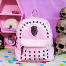 Load image into Gallery viewer, The GothX LIMITED EDITION Pastel Pink Skull Head Small Studs Vegan Mini Backpack on a pastel purple background with skulls and pastel pink coffin shelf in the background. The bag is facing forward to highlight the dark grey metal stud detailing, diamanté effect skull, front zip pocket, two side slip pockets and top handle.
