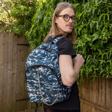 Load image into Gallery viewer, Jack is stood in a garden area modelling the pixel camouflage camo vegan backpack. The bag is facing towards the camera to highlight the front camo print, two front zip pockets, two elastic side pockets, detachable silver chain and top handle.
