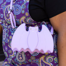 Load image into Gallery viewer, The GothX Pastel Lilac Purple Bat Vegan Shoulder Bag being modelled on a shoulder of someone wearing Run &amp; Fly purple paisley pinafore. The vegan leather pastel bag is facing forward to highlight the embroidered detailing and crystal eyes.
