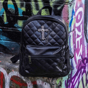 The GothX quilted cross vegan mini backpack sat outside on an alternative style colourful graffiti wall. The vegan leather bag is sat facing forward to highlight the quilted stitched front with a silver metal cross chain centre emblem, the front zip pocket, two side slip pockets, main top zip compartment and top handle.