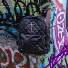Load image into Gallery viewer, The GothX quilted cross vegan mini backpack sat outside on an alternative style colourful graffiti wall. The vegan leather bag is sat facing forward to highlight the quilted stitched front with a silver metal cross chain centre emblem, the front zip pocket, two side slip pockets, main top zip compartment and top handle.
