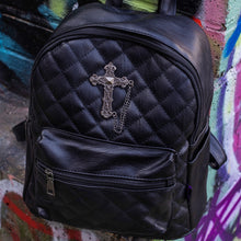 Load image into Gallery viewer, The GothX quilted cross vegan mini backpack sat outside on an alternative style colourful graffiti wall. The vegan leather bag is sat facing forward to highlight the quilted stitched front with a silver metal cross chain centre emblem, the front zip pocket, two side slip pockets, main top zip compartment and top handle.
