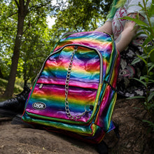 Load image into Gallery viewer, The CHOK rainbow holographic vegan shown next to a green haired model sat on a tree trunk. The bag has a rainbow melt holographic print with a detachable silver chain and CHOK logo. The bag is facing forward to highlight the front rainbow zip pockets, the main double rainbow zip compartment, two side elasticated pockets with a detachable decorative silver chain.
