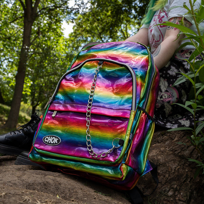 The CHOK rainbow holographic vegan shown next to a green haired model sat on a tree trunk. The bag has a rainbow melt holographic print with a detachable silver chain and CHOK logo. The bag is facing forward to highlight the front rainbow zip pockets, the main double rainbow zip compartment, two side elasticated pockets with a detachable decorative silver chain.